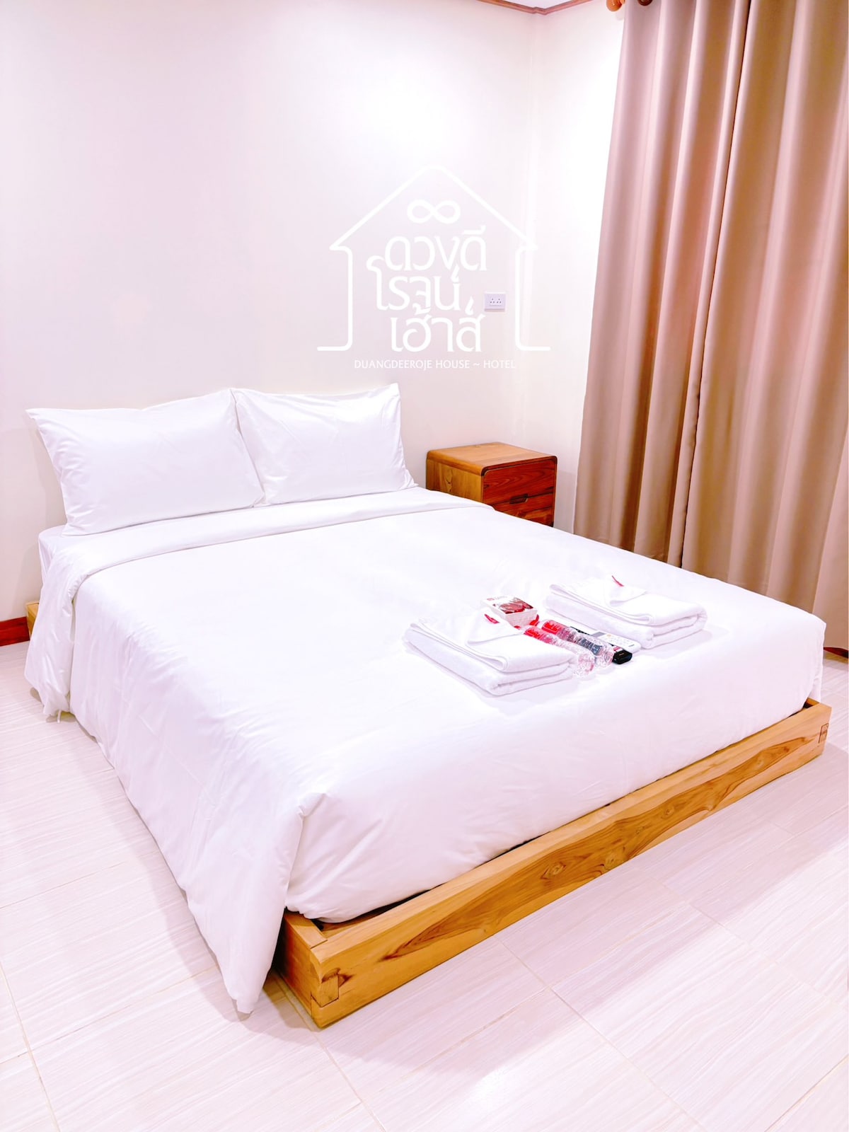 A bright sleeping area features a spacious bed with crisp white linens and two fluffy pillows. A wooden bedside table is positioned next to the bed, and light beige curtains frame a large window, allowing soft natural light to fill the room.