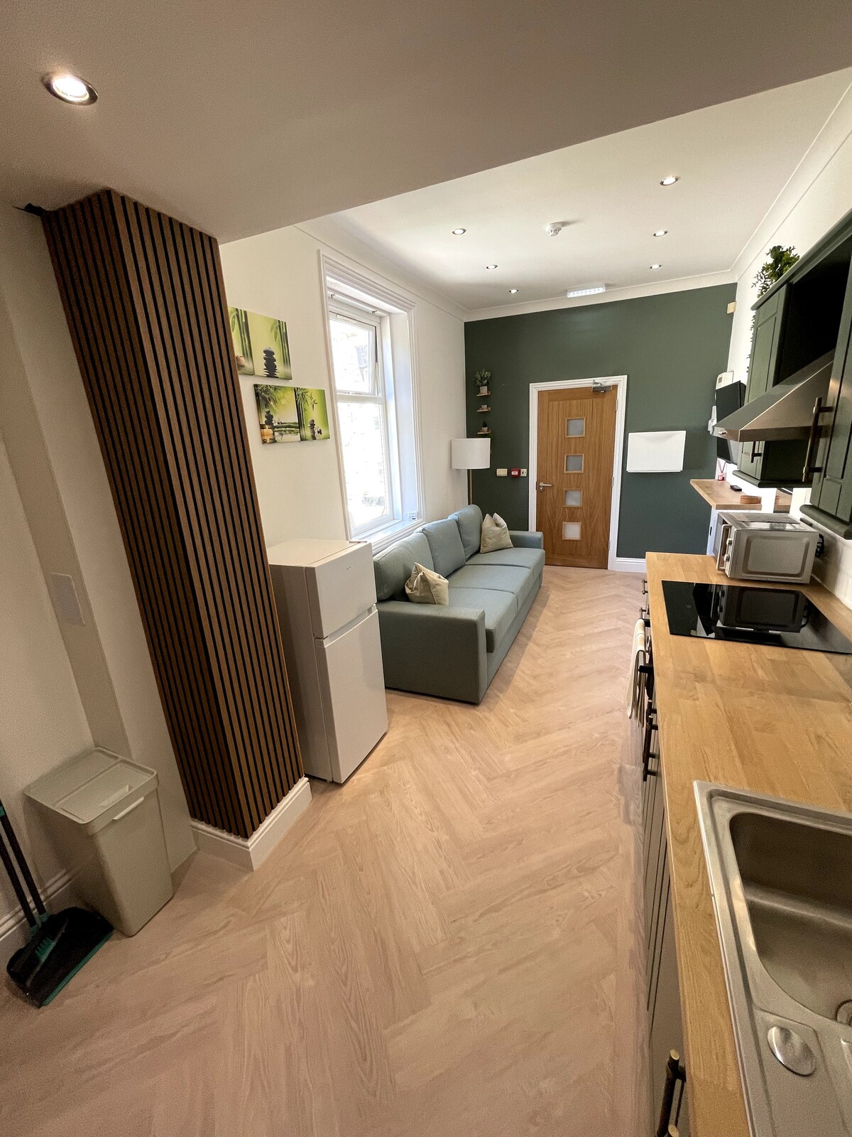 A compact kitchen area is featured, equipped with modern appliances including a refrigerator and microwave. A cozy sofa is placed against the wall, offering a comfortable seating option. Natural light streams in through the window, enhancing the neutral-toned decor.