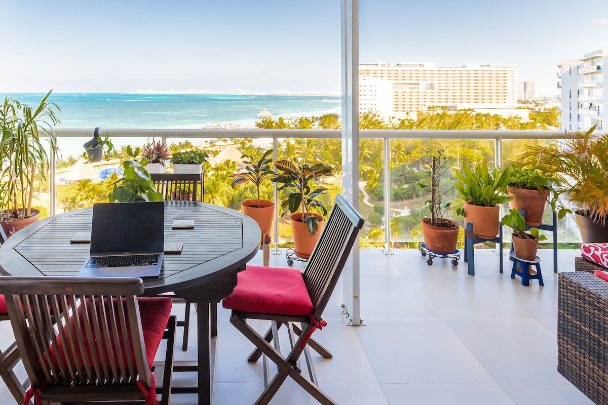 A spacious terrace features a round wooden table with a laptop, surrounded by chairs and vibrant potted plants. The serene view of the ocean is clearly visible, with the coastal landscape providing a tranquil backdrop. Natural light fills the area, enhancing the inviting atmosphere.