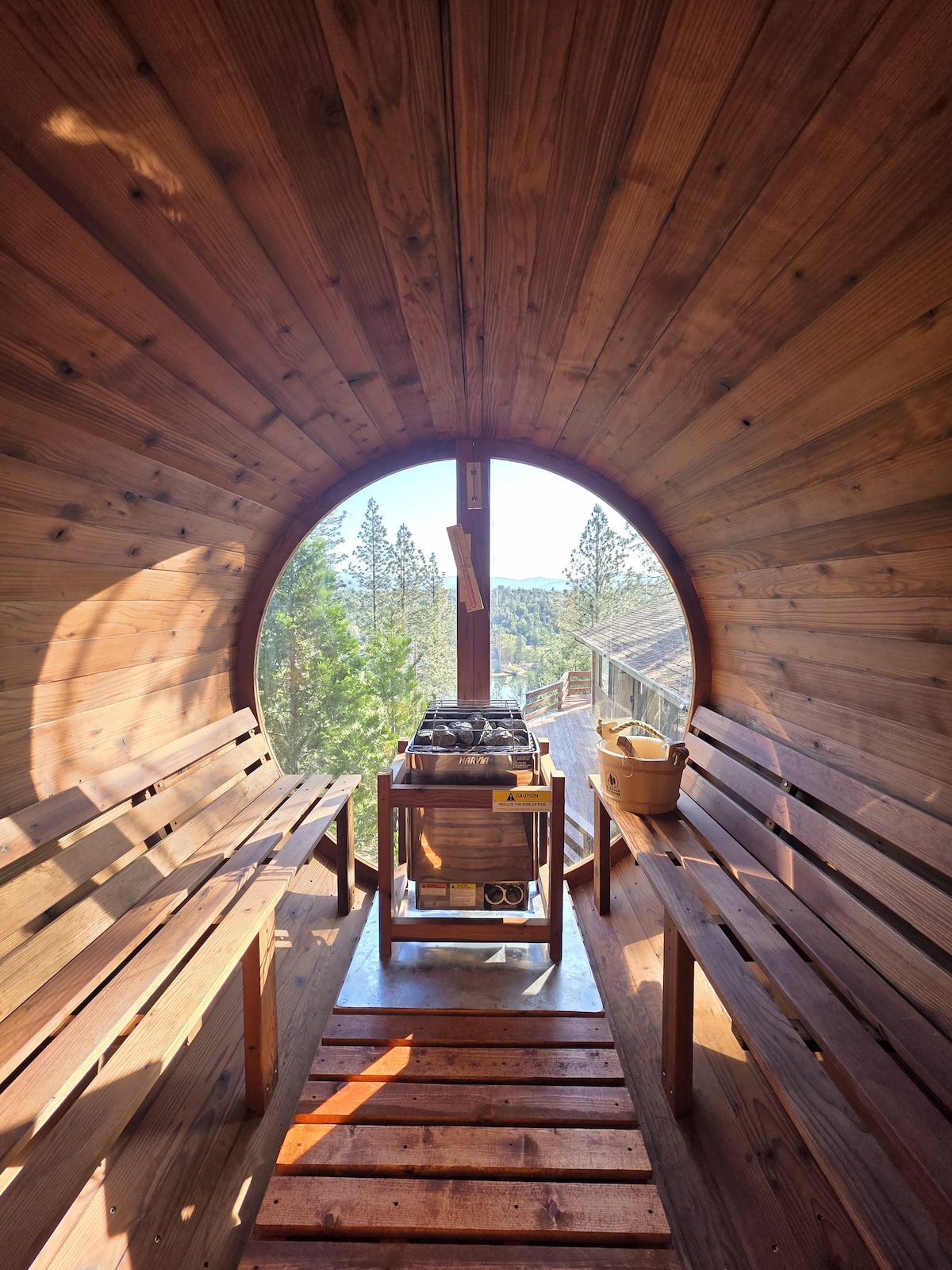 A panoramic barrel sauna features warm wooden interior, with benches on either side and a heater in the middle. The circular window offers expansive views of the surrounding trees, allowing natural light to illuminate the space.