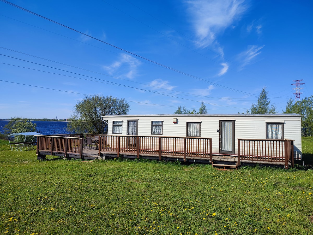 A large camper is situated on a grassy area near the Daugava river reservoir. Wooden decking extends across the front, with steps leading up to the entrance. Windows with sheer curtains allow light to enter, and a swing can be seen in the background.
