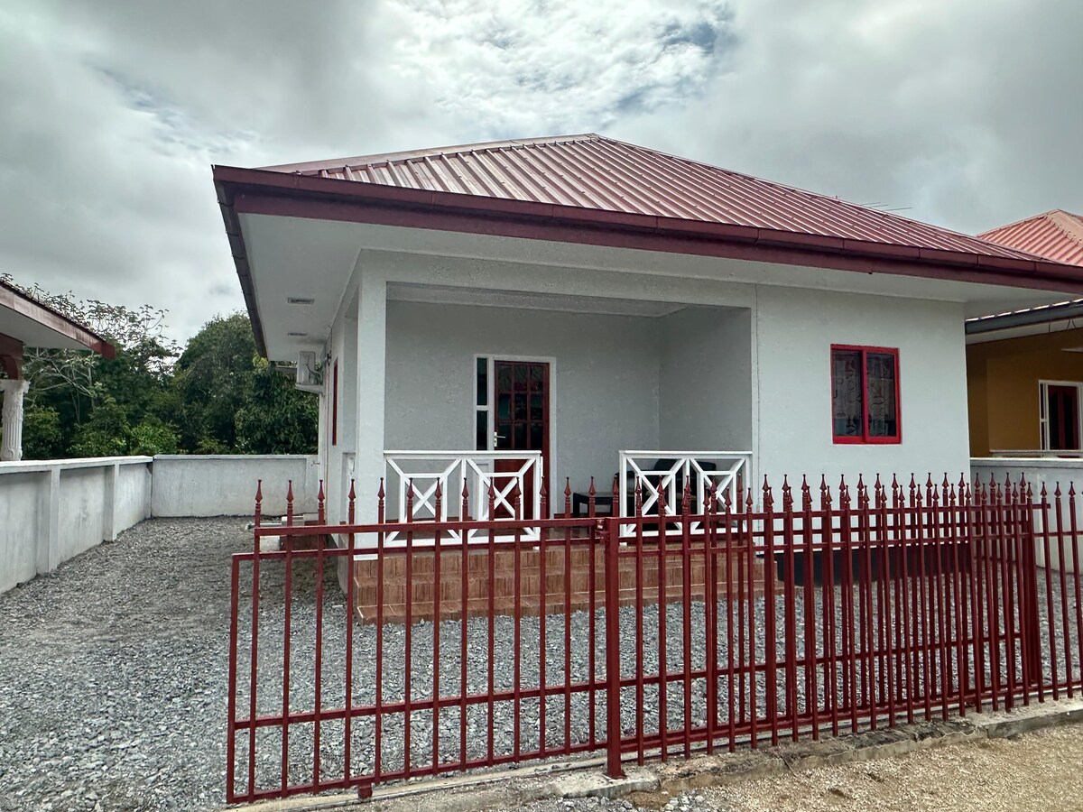 A single-story house is presented with a red metal roof and white walls. A porch area is featured at the front, with a railing and two chairs visible. Gravel surrounds the house, and a low fence encloses the yard. Gray clouds are observed in the sky.