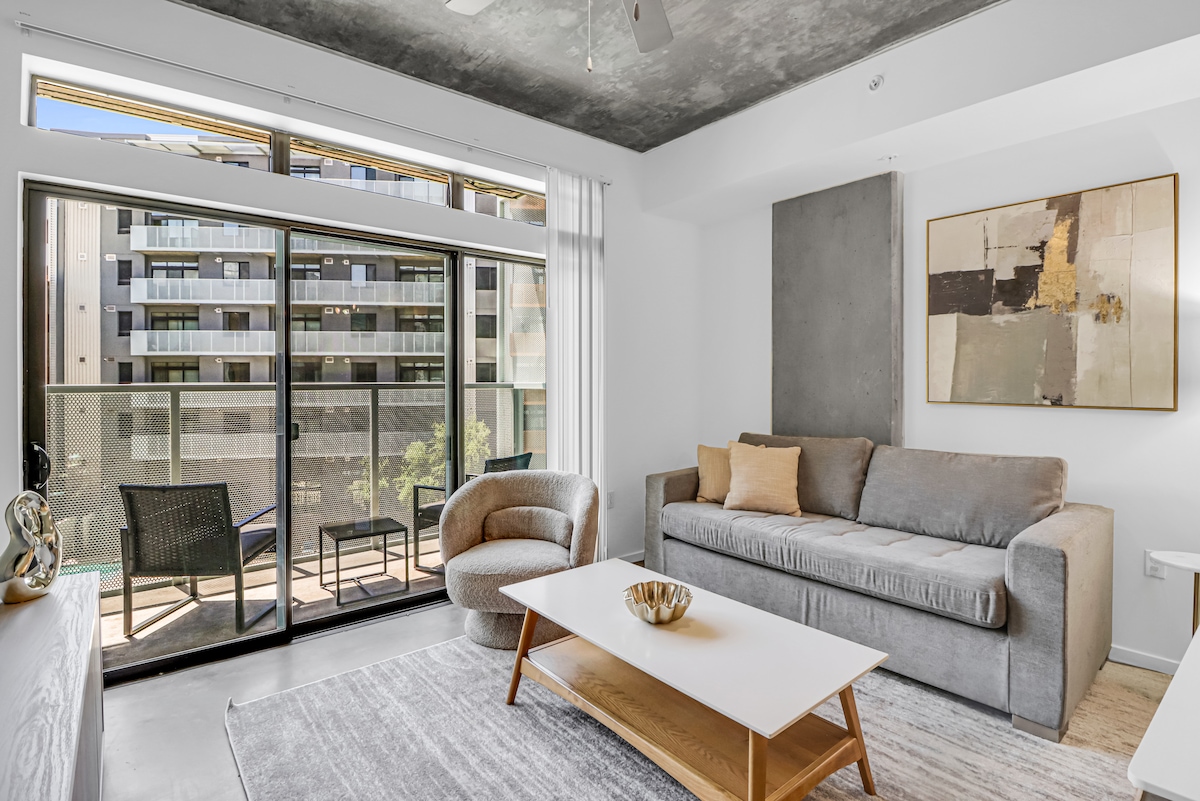 A spacious living area features a cozy sofa and an inviting accent chair. Large glass doors open to a balcony, allowing natural light to fill the room. A modern coffee table is centered on a soft area rug, complemented by contemporary wall art.
