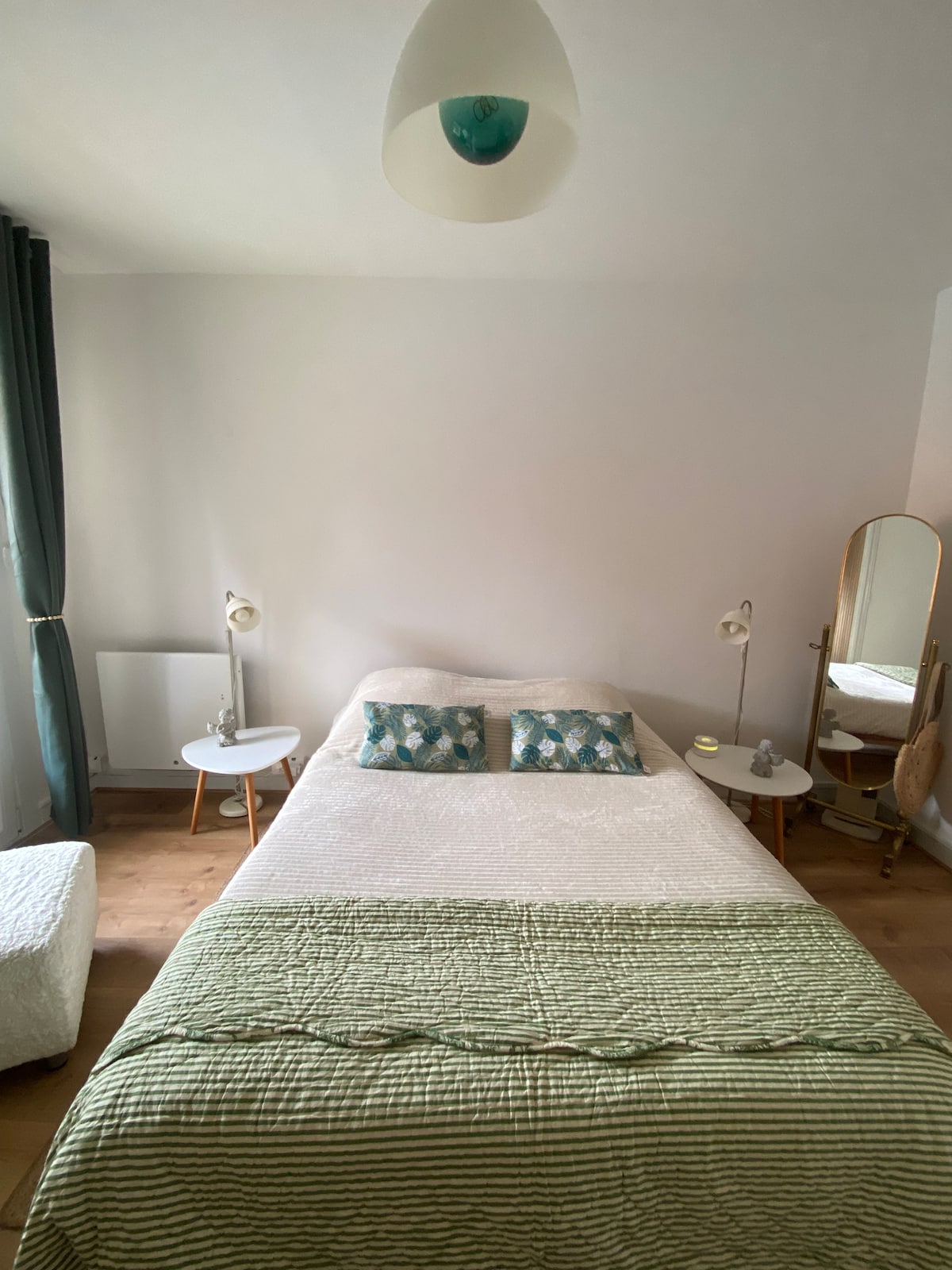 A cozy bedroom is presented with a spacious bed adorned with textured bedding and two decorative pillows. Soft light is provided by the bedside lamps. A mirror and small table enhance the minimalist decor, while natural light filters through the curtains.