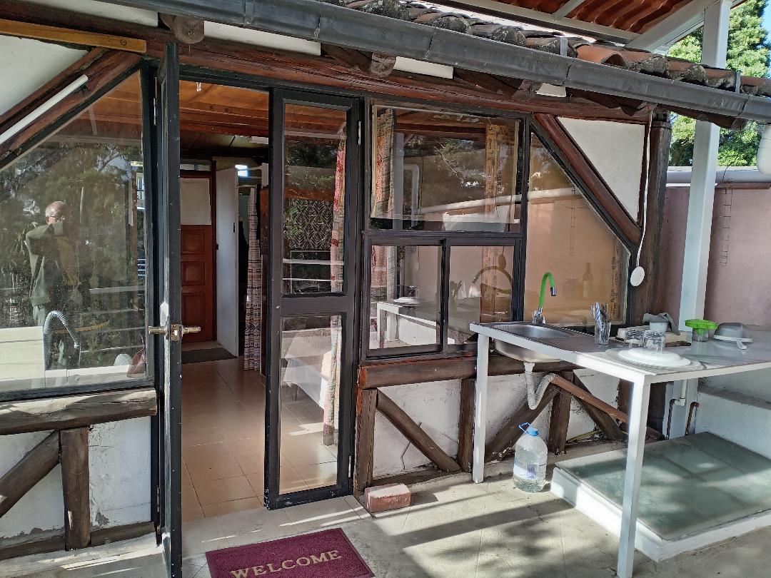 An outdoor kitchen area is shown with a countertop and a sink under a sloped roof. Large windows allow natural light in, while a welcome mat is placed on the tiled floor. The entryway is framed by wooden beams, leading into the living space.