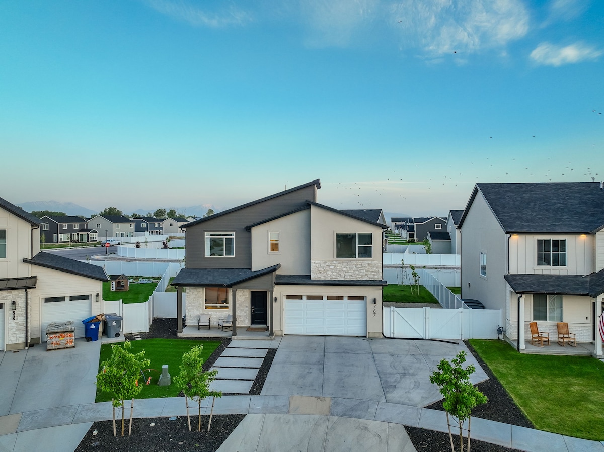 A modern two-story home is displayed, featuring a structured façade with a mix of stone and siding. The driveway is flanked by a neatly manicured lawn, while a spacious front porch leads to the entrance. White fencing surrounds the yard, contributing to a sense of privacy.