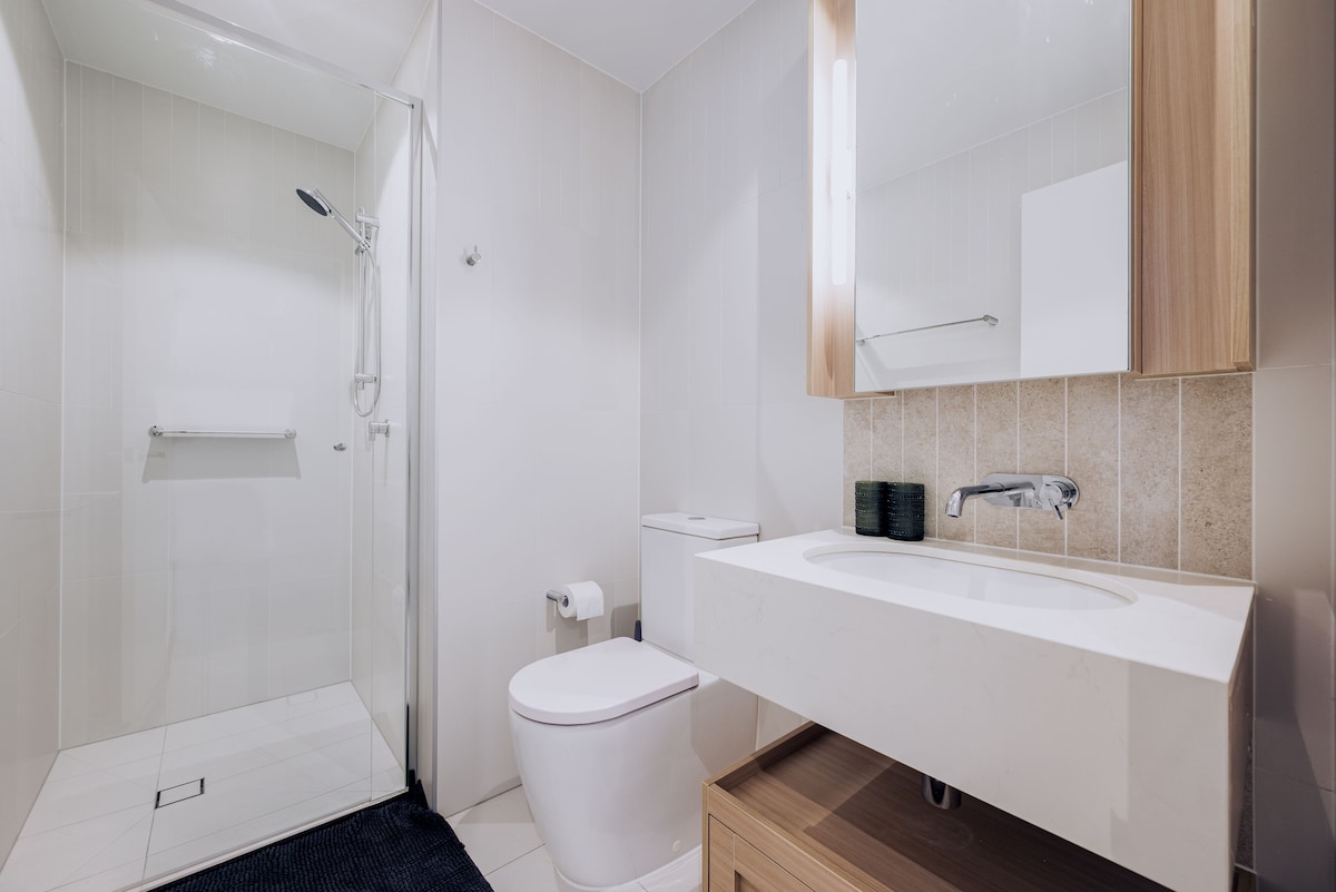 A modern bathroom features a spacious walk-in shower with a glass enclosure and a sleek rain showerhead. The elegant vanity has a minimalist design with a large mirror above. A soft navy mat is placed on the floor, complementing the understated neutral tones of the room.
