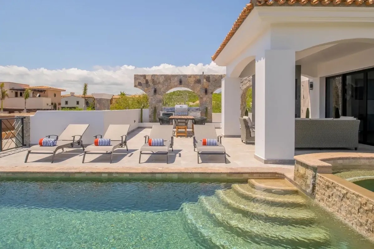A spacious terrace features a heated pool with gently sloping steps. Six lounge chairs are arranged around the pool, and an outdoor dining area is visible under a shaded archway. The surrounding landscape offers a blend of natural and built environments.
