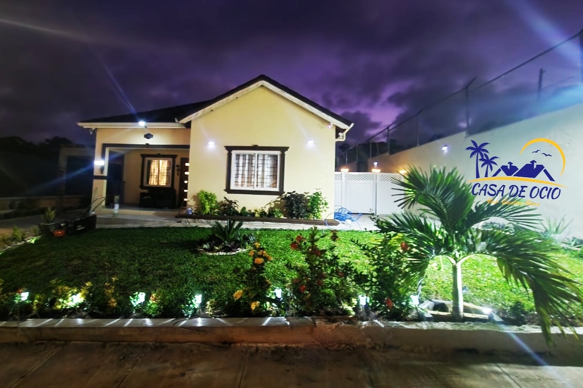 The exterior of a charming yellow house is showcased, with a well-manicured lawn featuring colorful flower beds. Soft lighting illuminates the walkway, and a decorative palm plant is visible in the foreground. The evening sky adds a serene backdrop to the scene.