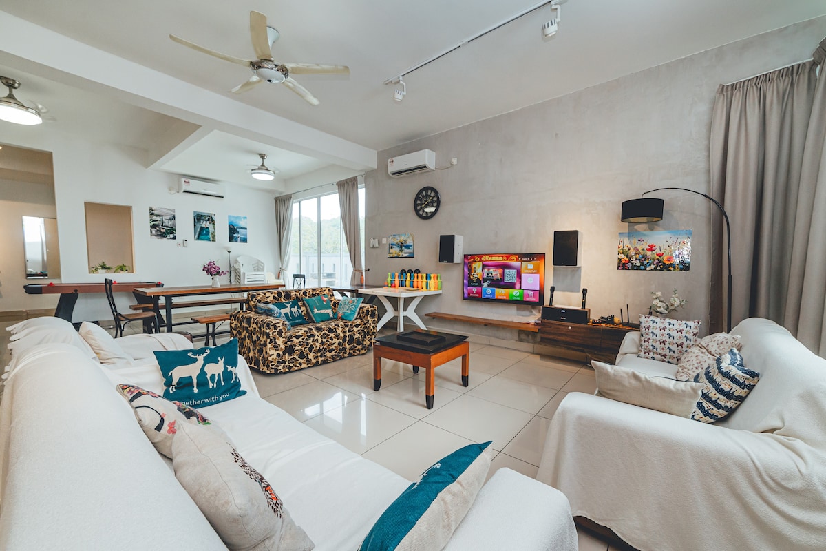 The spacious living area features a light-colored tiled floor and offers various seating options. Two patterned sofas and a wooden coffee table are positioned around a flat-screen TV, which is mounted on the wall. Natural light enters through large windows, complemented by soft curtains.