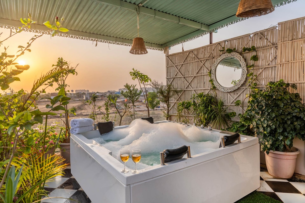 A private rooftop jacuzzi is showcased, surrounded by lush greenery and plants. Two glasses of wine sit on the edge, complementing a bubbly atmosphere. The sunset casts a warm glow over the scene, creating a serene setting for relaxation.