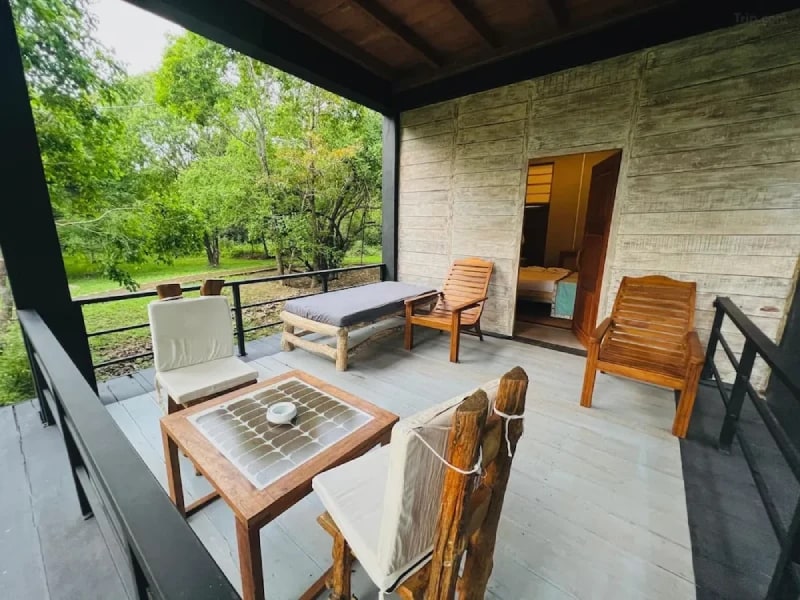A private terrace is furnished with a wooden table and chairs, complemented by a lounge chair. Natural surroundings are visible beyond the railing, enhancing the connection to nature. An entrance leads to an interior space featuring a sleeping area, framed by a rustic wall texture.