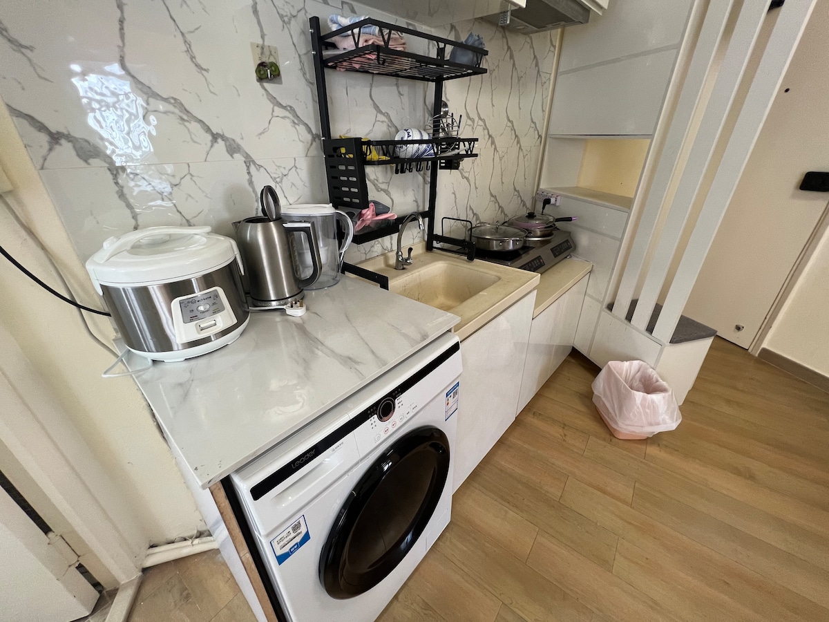 A compact kitchen area features a marble-patterned backsplash and a variety of appliances, including a kettle and a rice cooker. An integrated cooking space is equipped with a gas stove, while a washing machine is positioned beside the sink.