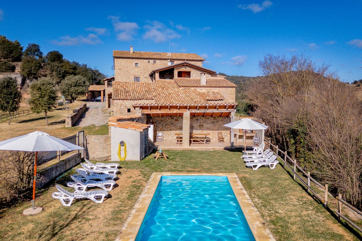 The exterior of the masía is showcased, featuring a swimming pool with clear blue water surrounded by lounge chairs. Sun umbrellas offer shade, and a grassy area is visible alongside the pool. The rustic stone structure is set against a backdrop of rolling hills and trees.