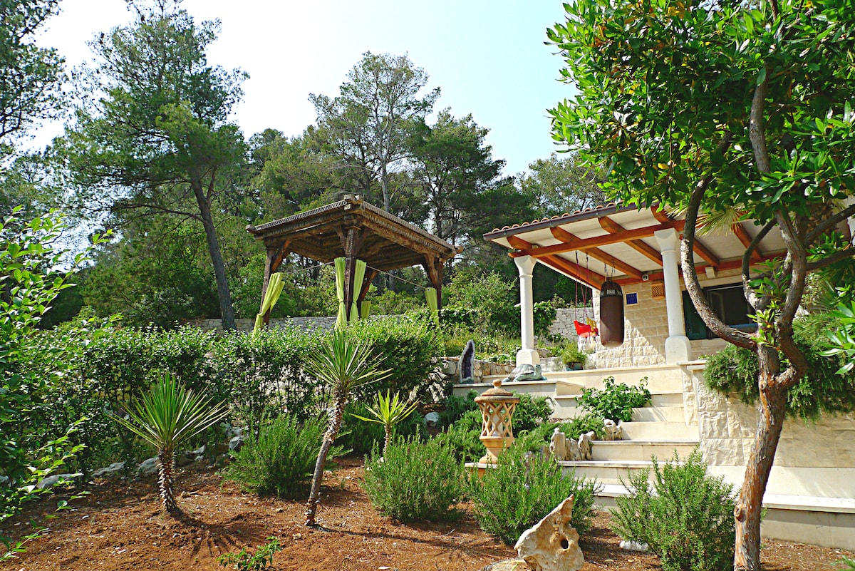 A shaded pergola is positioned among lush greenery, providing a serene outdoor space. Surrounding vegetation includes various Mediterranean plants and stone sculptures, contributing to a tranquil atmosphere. The villa's exterior features natural stone walls and wooden accents, blending harmoniously with the landscape.
