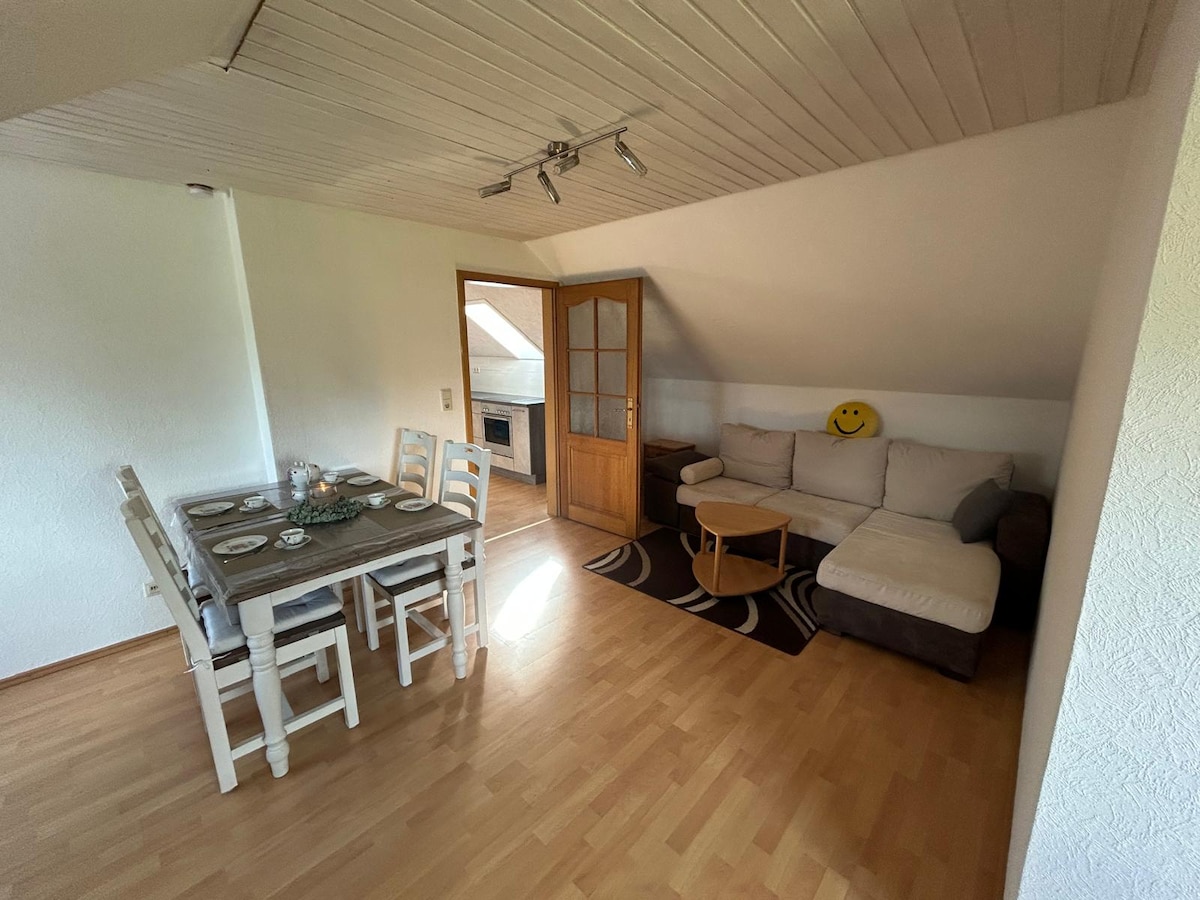 A light-filled living area features a comfortable sectional sofa alongside a small coffee table. A dining table with seating for six is placed near the windows. A door leads to the kitchen area, while warm wooden flooring complements the overall space.