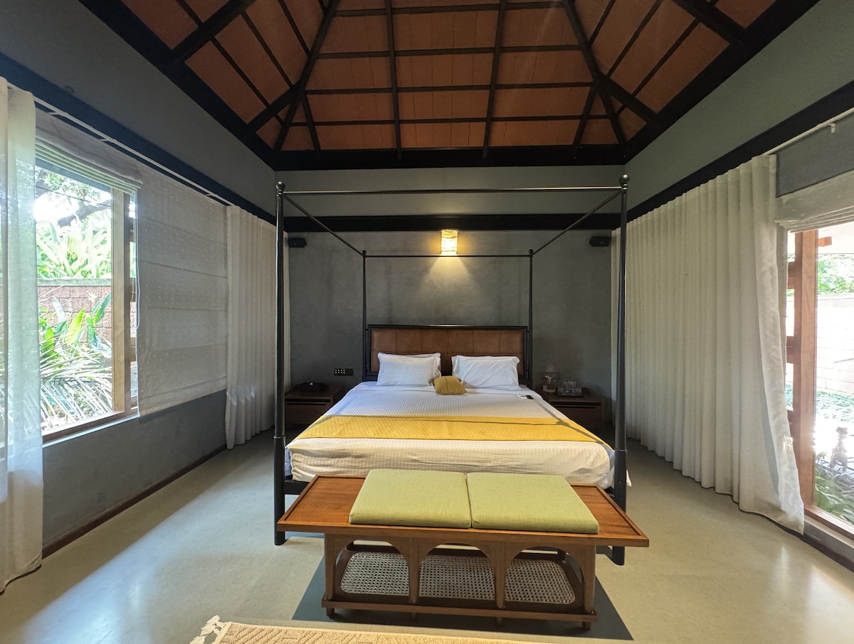 A spacious bedroom features a four-poster bed adorned with crisp white linens, complemented by a wooden bench in front. Soft curtains frame the large windows, allowing natural light to fill the room. The high ceiling enhances the open and airy feel of the space.