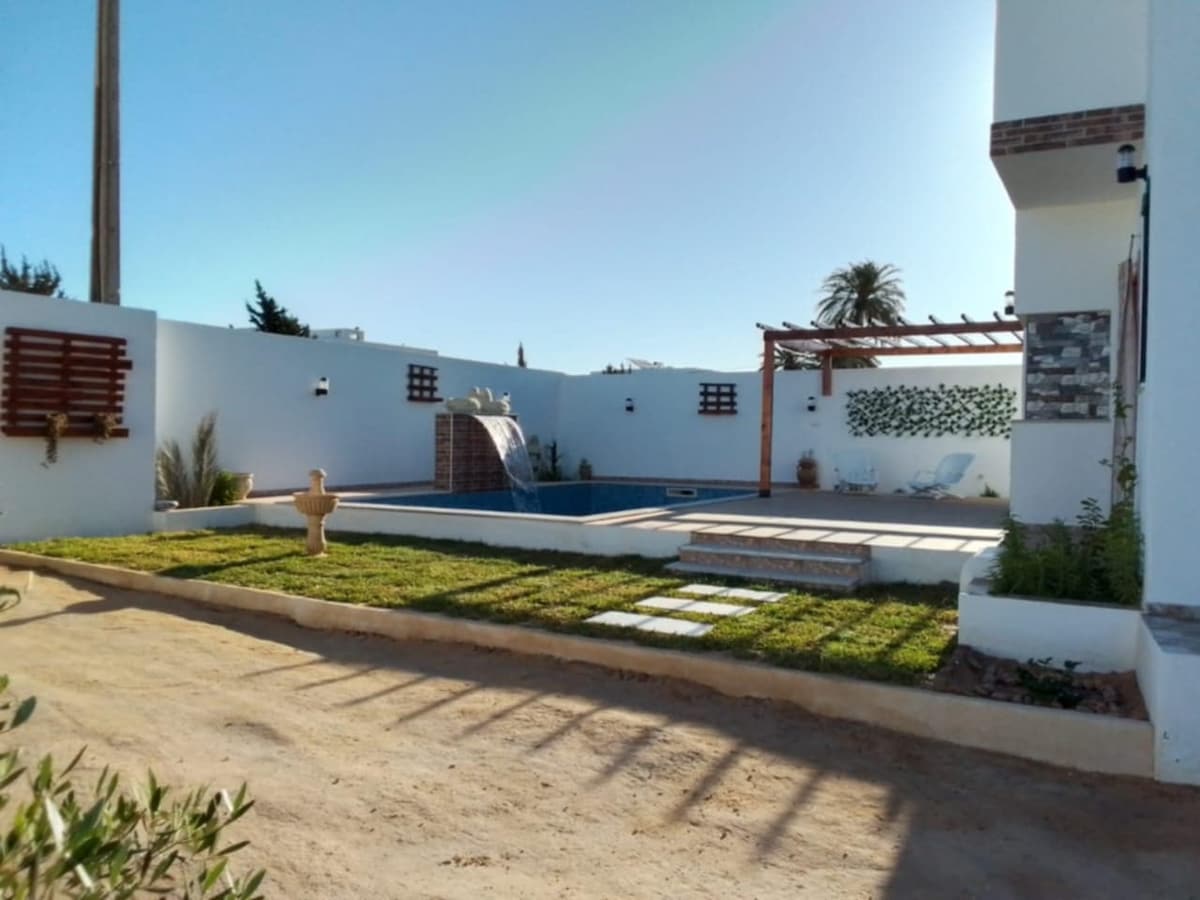 A private pool is surrounded by well-maintained grass and stone pathways. A decorative fountain adds an elegant touch to the outdoor space, while a pergola and comfortable seating are visible in the background, offering a serene environment for relaxation.