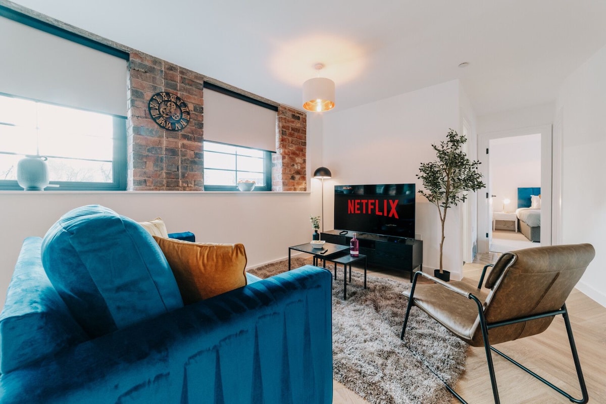 An open-plan lounge features modern décor, highlighted by a blue sofa and brown accent chair. A large TV displays the Netflix logo. Natural light enters through two windows adorned with light-colored blinds, creating a comfortable and inviting living space.