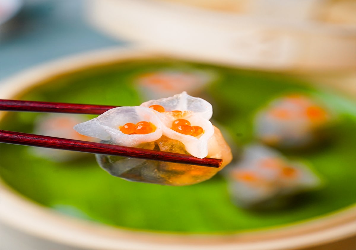 A close-up view of a delicately crafted dim sum dish is presented. The dumplings are shaped like flowers, showcasing translucent skin and orange fish roe on top. A bamboo steamer is visible in the background, resting on a green leaf.