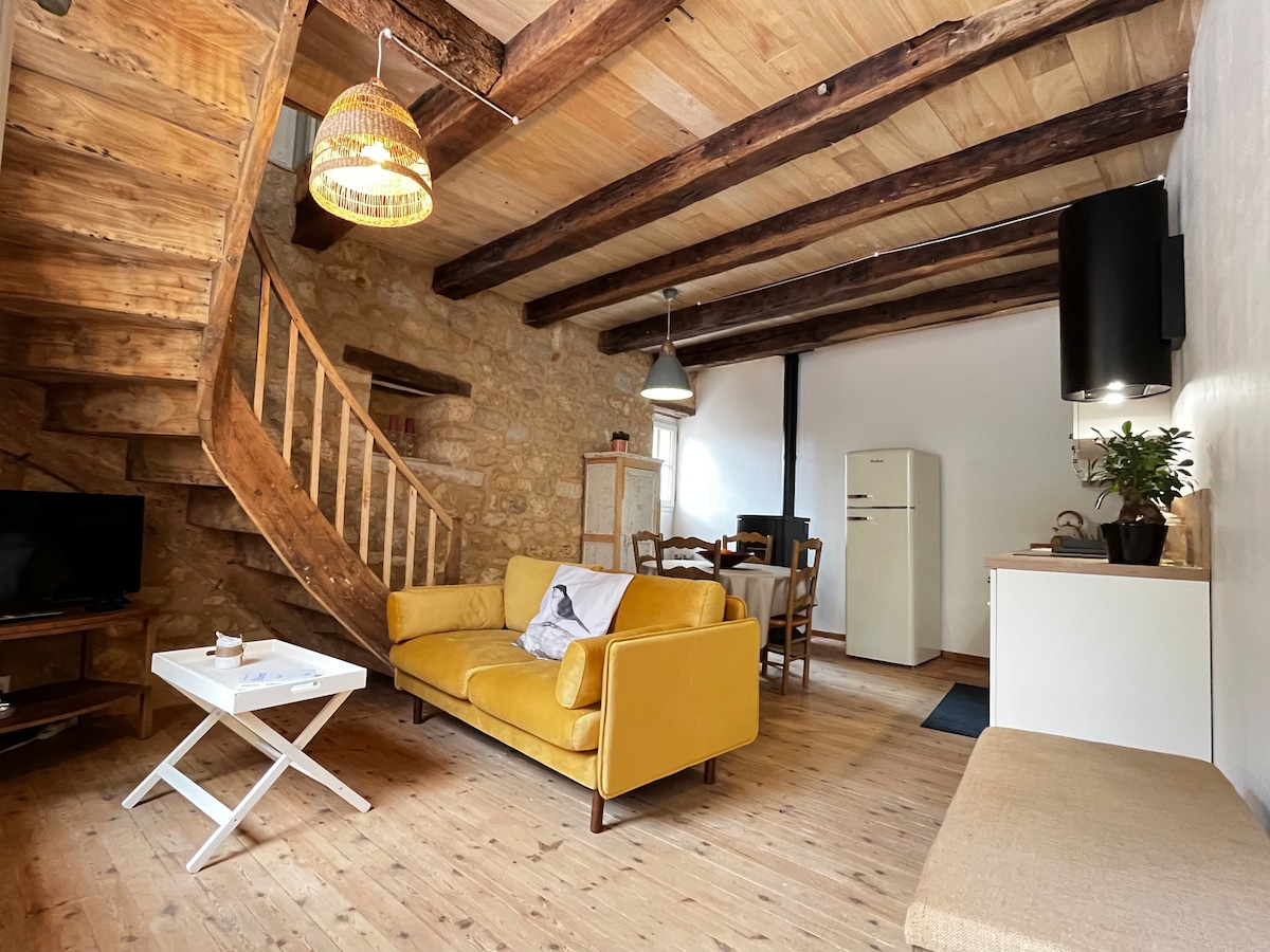The living area features exposed wooden beams and a warm wood floor. A yellow couch sits beside a small coffee table, with a TV positioned on a stand to the left. A dining table with four chairs is visible, along with a white refrigerator and a minimalist kitchen area.