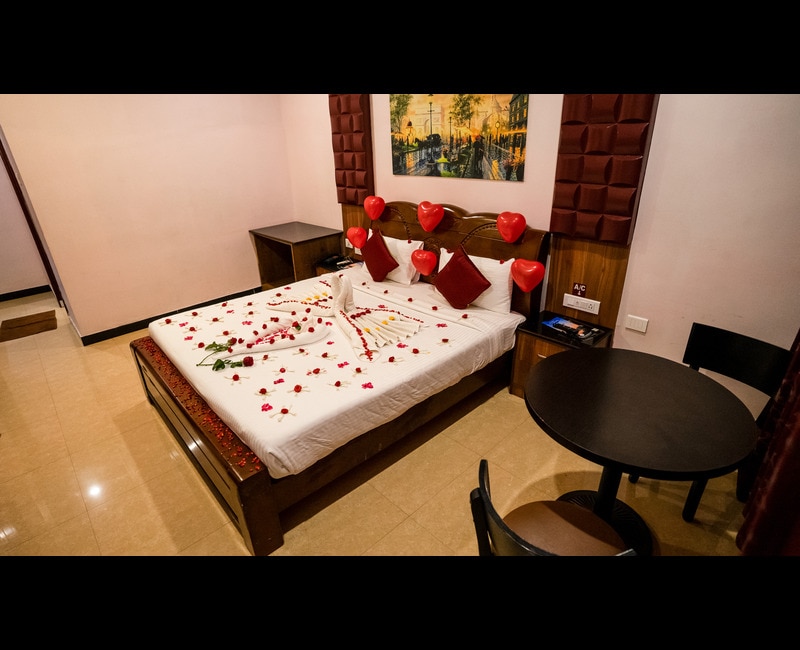 A spacious bedroom is presented with a neatly arranged bed adorned with heart-shaped balloons and rose petals. A small dining table and chair are positioned nearby, with a warm wall color and a decorative piece in the background adding to the room's charm.