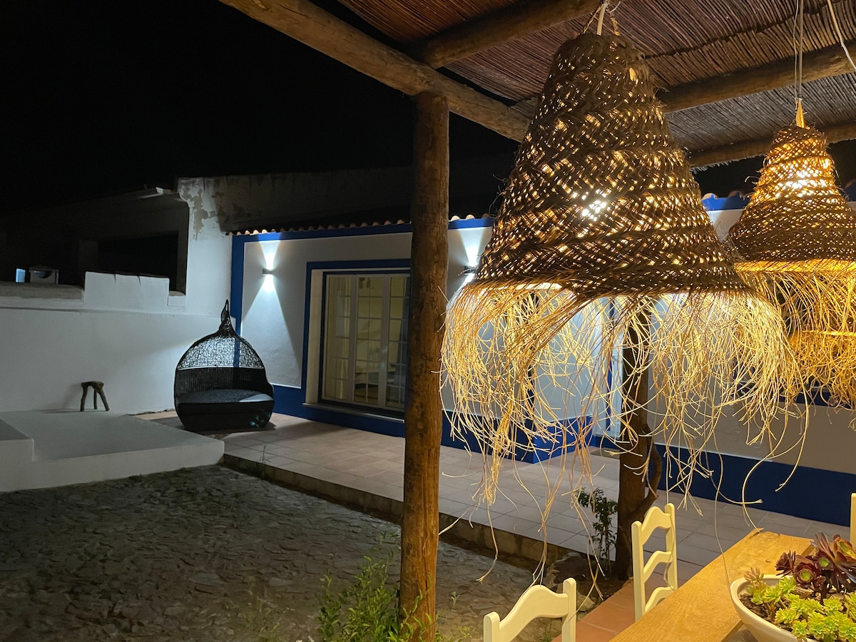An outdoor dining space is highlighted by two woven pendant lights, casting warm shadows. In the background, a charming area features a freestanding hanging chair and soft lighting reflecting off the white walls of the house, creating a serene evening ambiance.