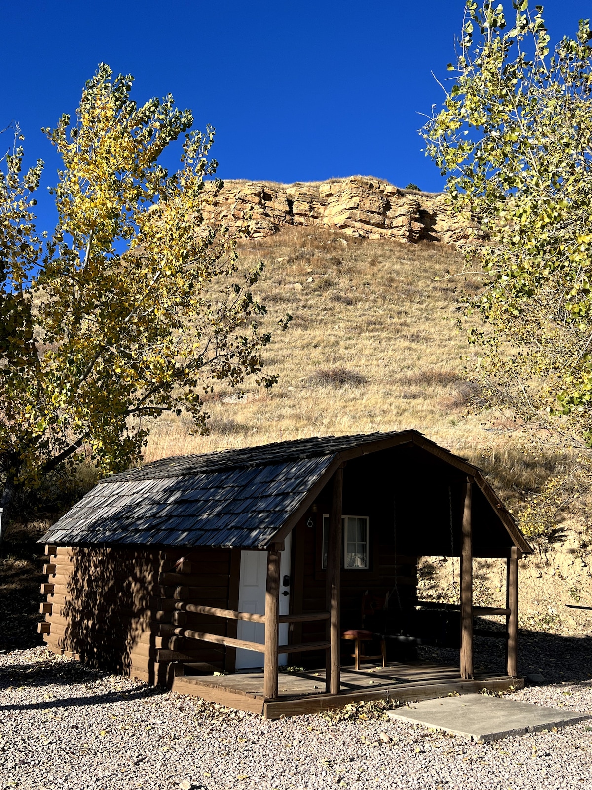 A rustic cabin sits at the base of a hillside, surrounded by trees displaying autumn leaves. The cabin's porch features a simple seating area, and a stone path leads to the entrance. A clear blue sky provides a bright backdrop for the natural landscape.