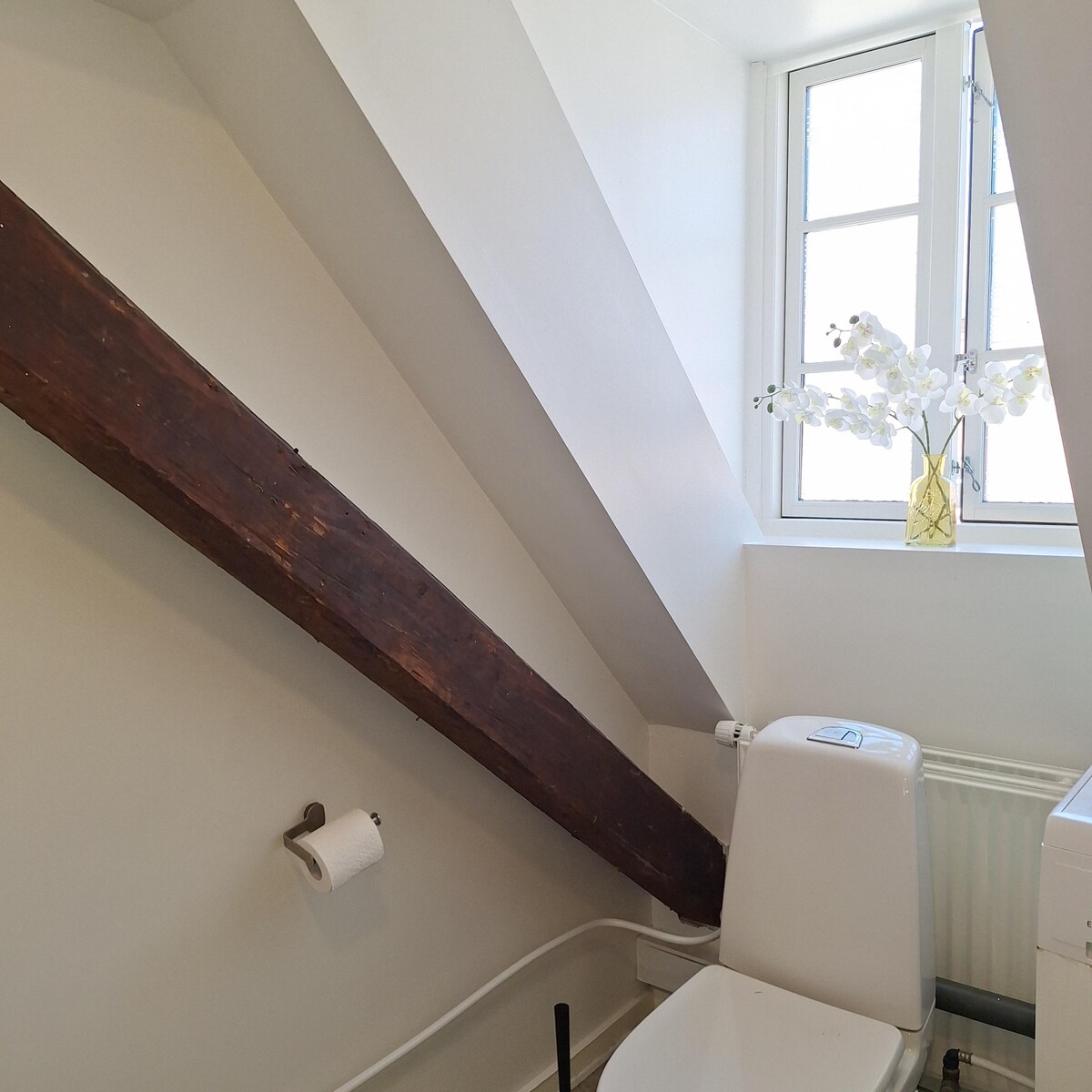 A small bathroom area features sloping walls with exposed wooden beams. A white toilet is positioned adjacent to a window adorned with a vase of orchids, providing a touch of decor. Soft, natural light enters through the window, illuminating the space.