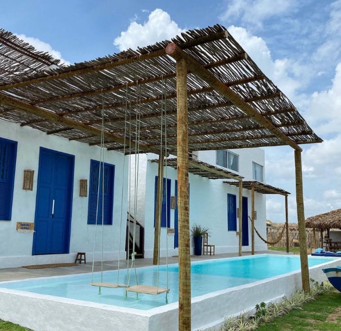 A restful outdoor area features a shaded wooden terrace, with a swing hanging above a refreshing pool. The white walls of the building contrast with the blue doors, creating a serene and inviting environment. Lush green landscaping borders the pool, enhancing the natural setting.