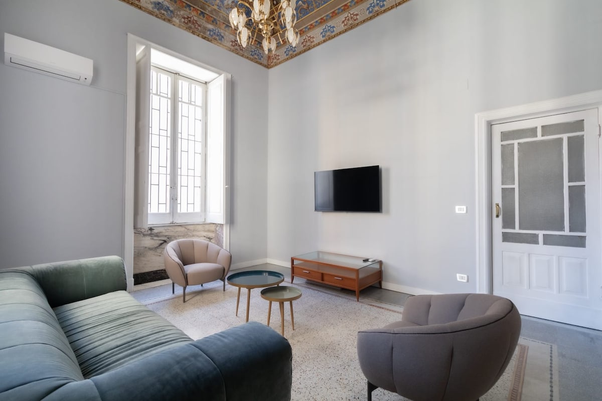 A well-lit living area features a modern sofa in soft fabric, accompanied by two elegant chairs. A small, round table sits in the middle. The room is adorned with a frescoed ceiling and large windows allowing natural light to enter, enhancing the space's comfort.