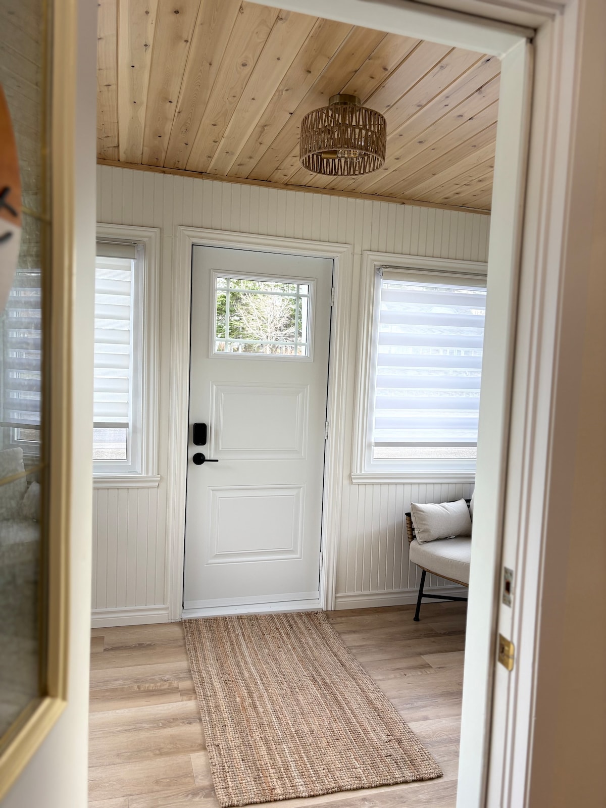 A bright entryway features a white door with a glass panel, framed by large windows with sheer shades. A woven rug lies on the wooden floor, leading to a cozy seating area with a minimalist chair. Natural wood paneling adorns the ceiling.