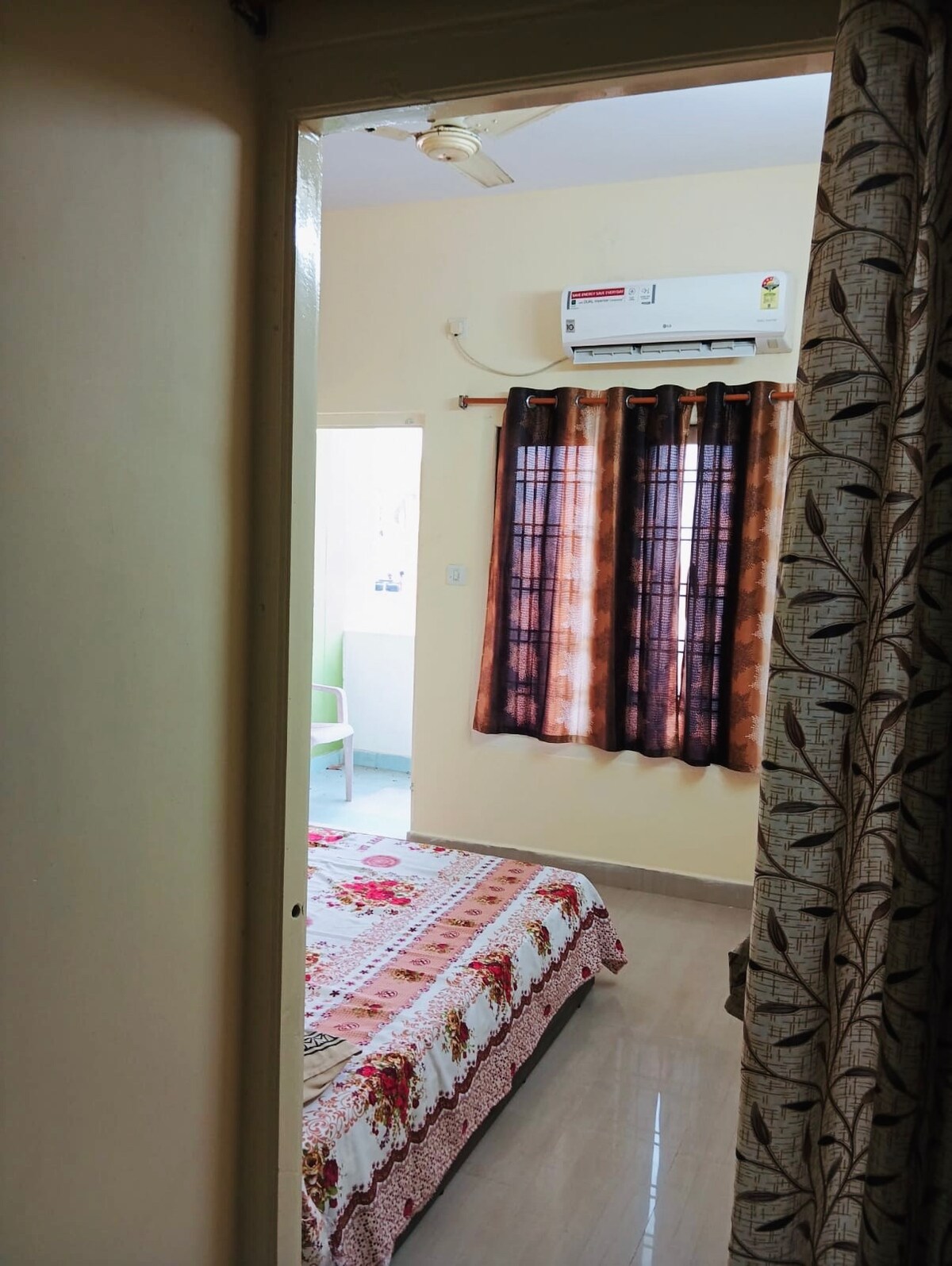 A glimpse of a cozy sleeping area is offered through an open door. The room features a spacious bed adorned with a floral blanket. Dark curtains frame a window that allows natural light to brighten the space, while an air conditioning unit is mounted above.