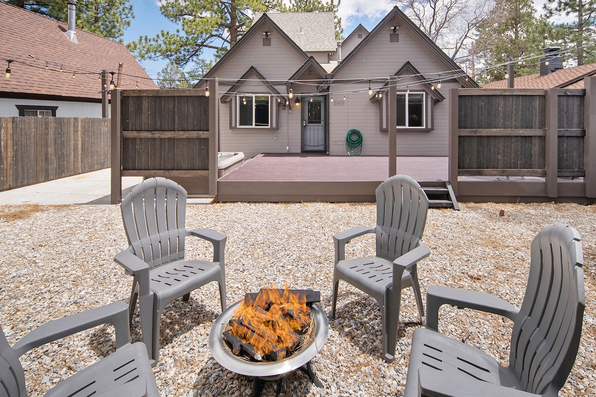 A gravel outdoor area features a fire pit surrounded by four gray chairs. The home's exterior is visible in the background, showcasing a spacious deck and string lights overhead. The fully fenced yard provides a secure and private setting.