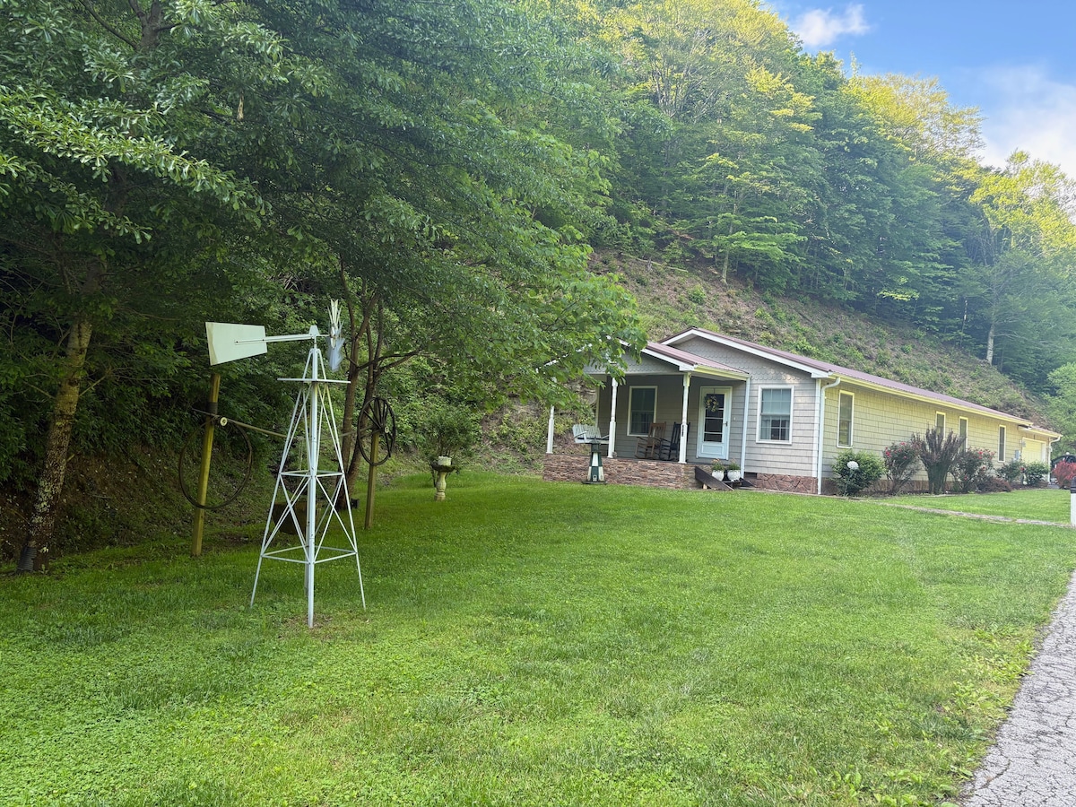 A pleasant exterior view of a single-story home surrounded by greenery. A windmill stands in the yard, complementing the natural landscape. The house features a porch and large windows, providing a welcoming appearance amid the lush hillside.