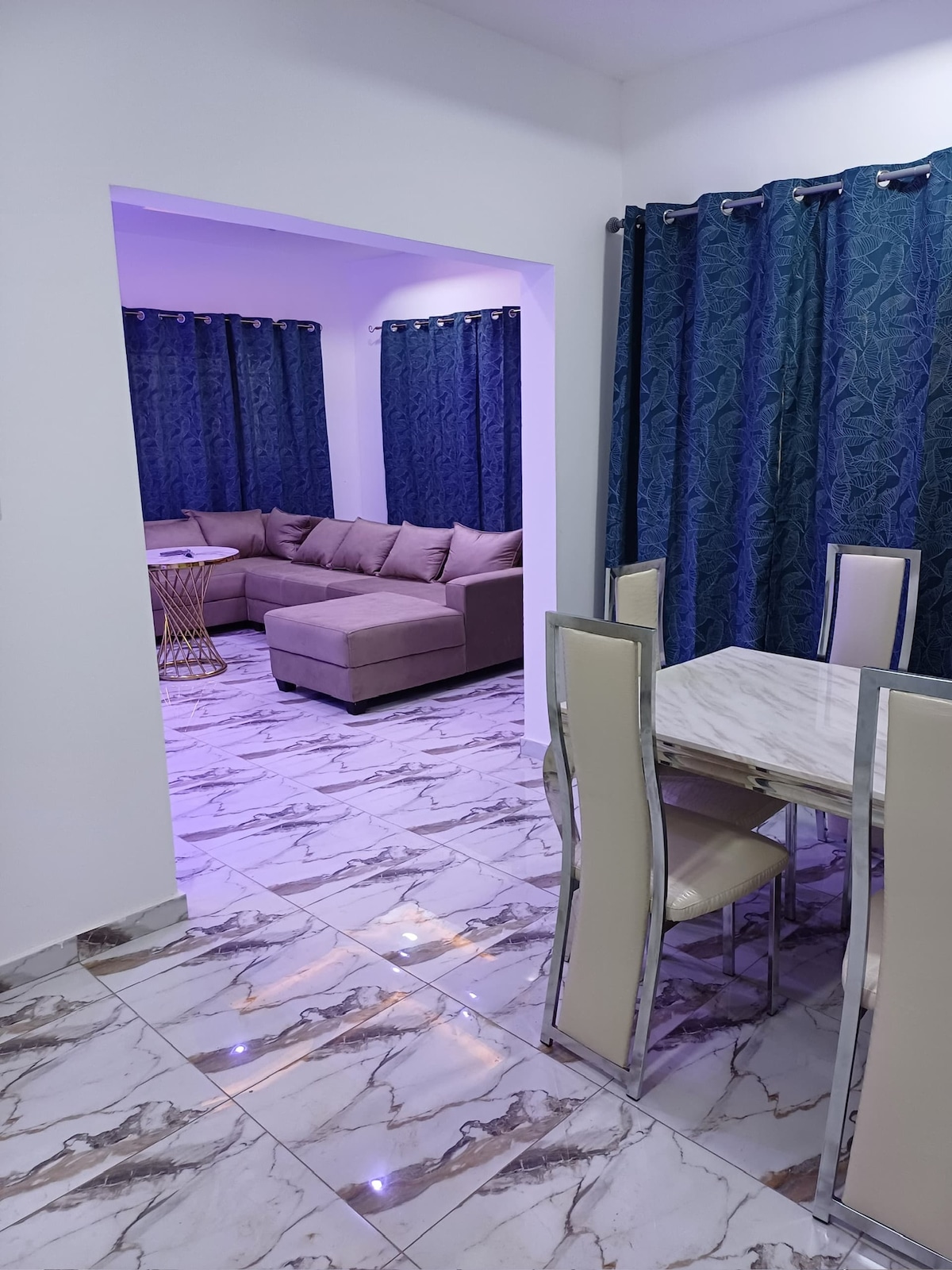 A modern dining area with a marble table and light-colored chairs is visible. The adjoining seating area is furnished with a spacious sectional couch, complemented by deep blue curtains. Ambient lighting adds a gentle glow to the space, enhancing its design and inviting feel.