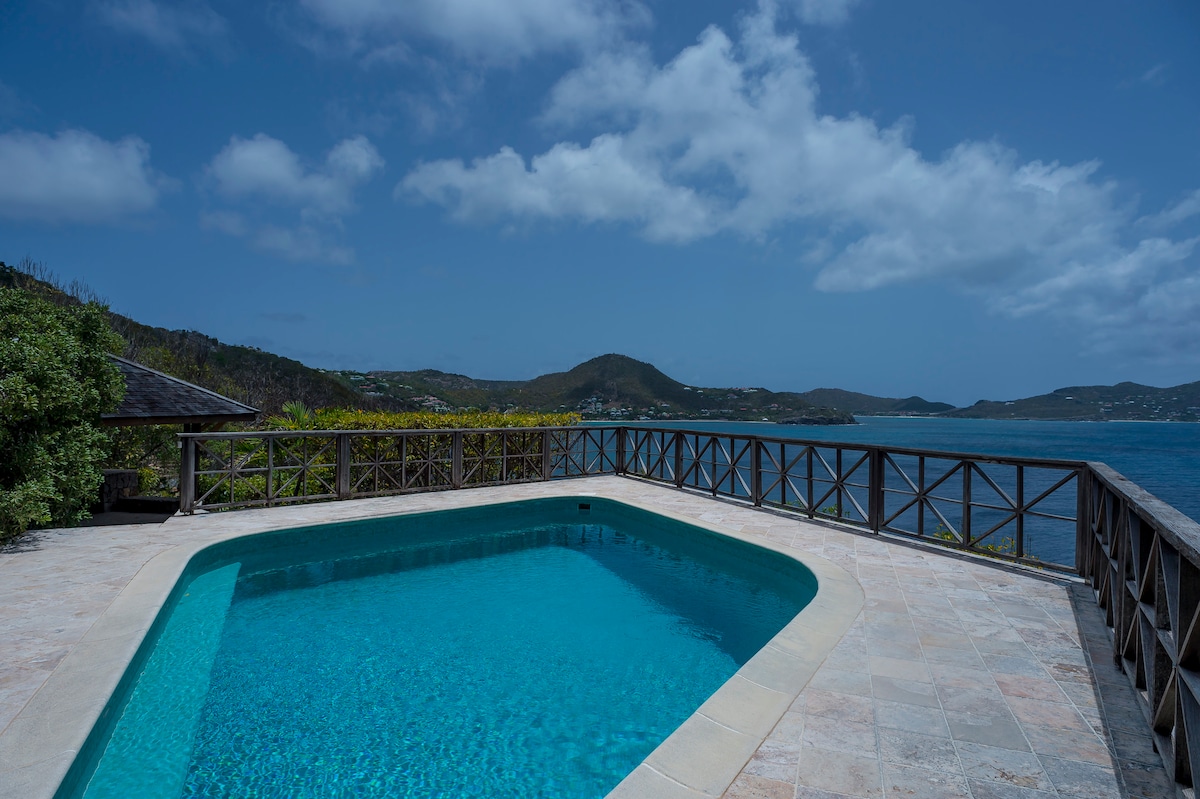 A serene pool area is captured, featuring a clear blue pool bordered by natural stone. Lush greenery and wooden railings frame the space, with a stunning ocean view visible in the background under a partly cloudy sky.