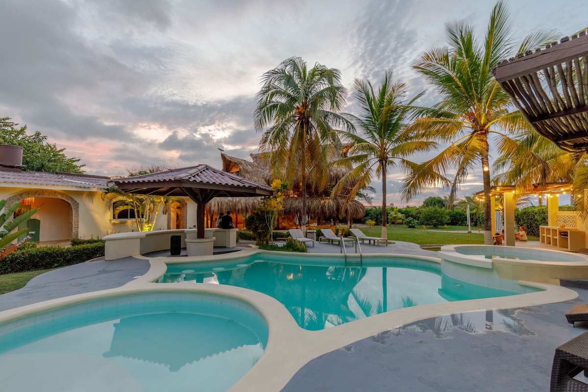 A tranquil pool area is surrounded by palm trees, with varying depths creating distinct swimming spaces. An outdoor bar structure offers shaded seating, while the sky transitions into soft hues at sunset, reflecting a serene atmosphere. The well-maintained grounds extend beyond the pool.
