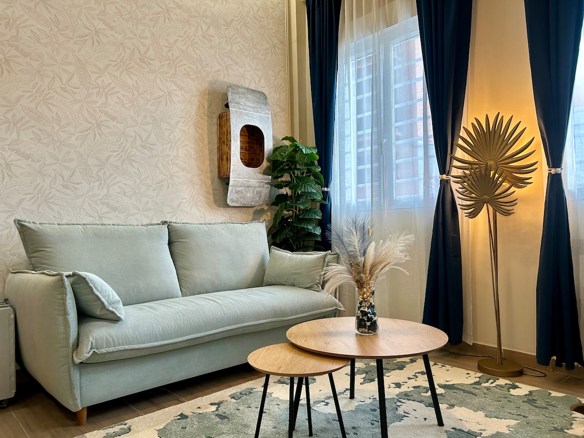 A soothing living area features a soft green sofa and two round wooden coffee tables. Natural light filters through sheer curtains, highlighting a decorative plant and a stylish floor lamp. A textured wall adds subtle depth to the space.