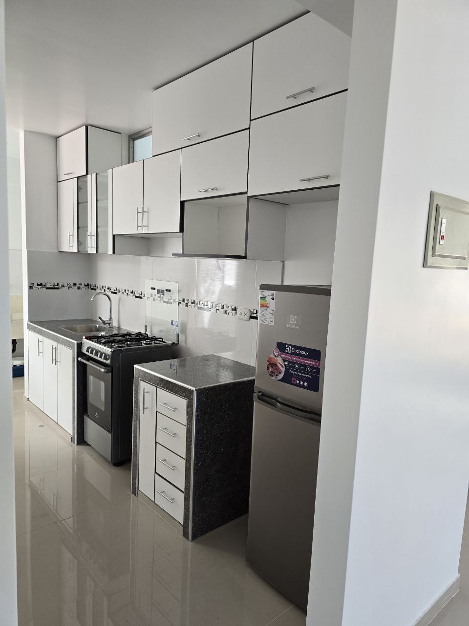 A modern kitchen is shown, featuring sleek white cabinets and a dark granite countertop. A gas stove and oven are positioned along one wall, while a silver refrigerator stands nearby. The design is clean and functional, with tiled backsplash adding subtle detail.