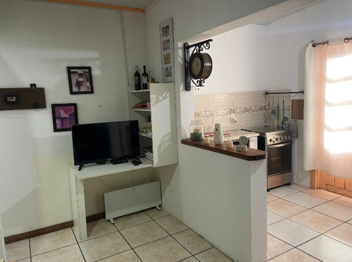 A spacious living area features a white entertainment unit with a television. The adjoining kitchen is visible, equipped with a stove and various kitchen supplies. Natural light filters in through a nearby window, illuminating the room's neutral-colored tiles and warm accents.
