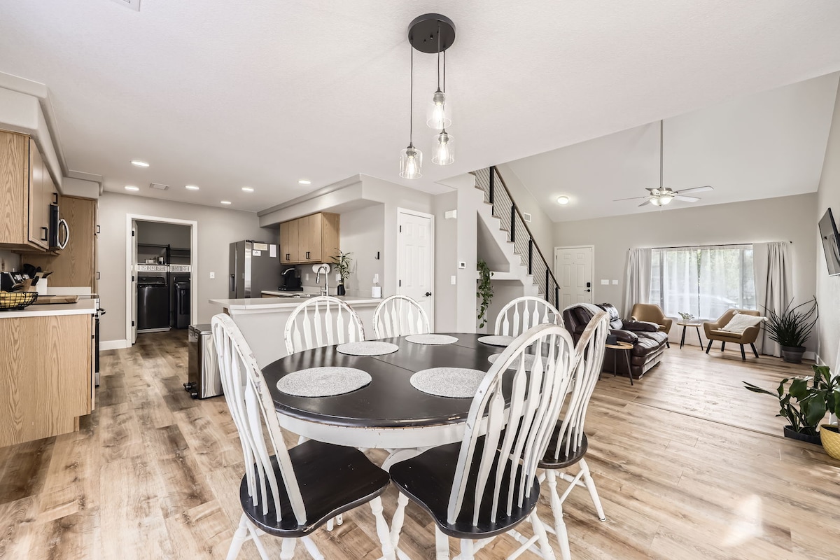 A modern dining area features a round black table surrounded by six white chairs. In the background, a spacious living room is visible, furnished with a brown couch, a ceiling fan, and large windows allowing natural light to illuminate the space.