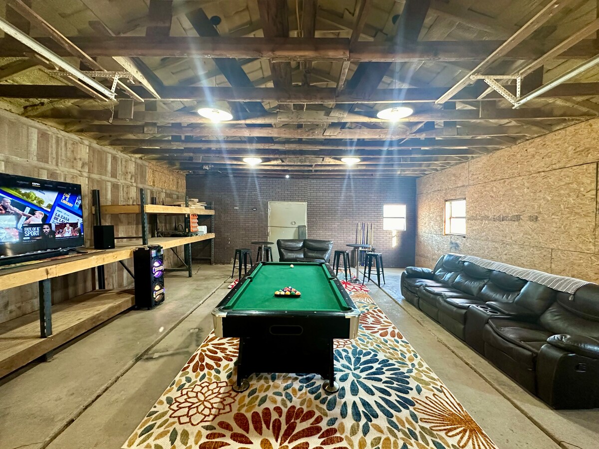 A cozy lounge area features a green pool table positioned centrally. Comfortable seating is arranged along the walls, while a large TV is mounted on the opposite side. Shelving units provide storage space, and a decorative rug adds a pop of color to the concrete floor.