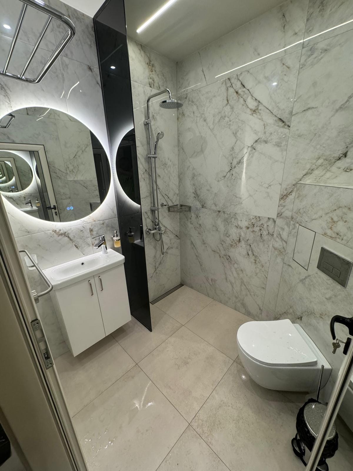 A modern bathroom features marble walls accented by circular mirrors. A sleek shower area is present, along with a compact sink and wall-mounted toilet. Soft lighting enhances the serene atmosphere, and a mobility resource is discreetly positioned nearby.