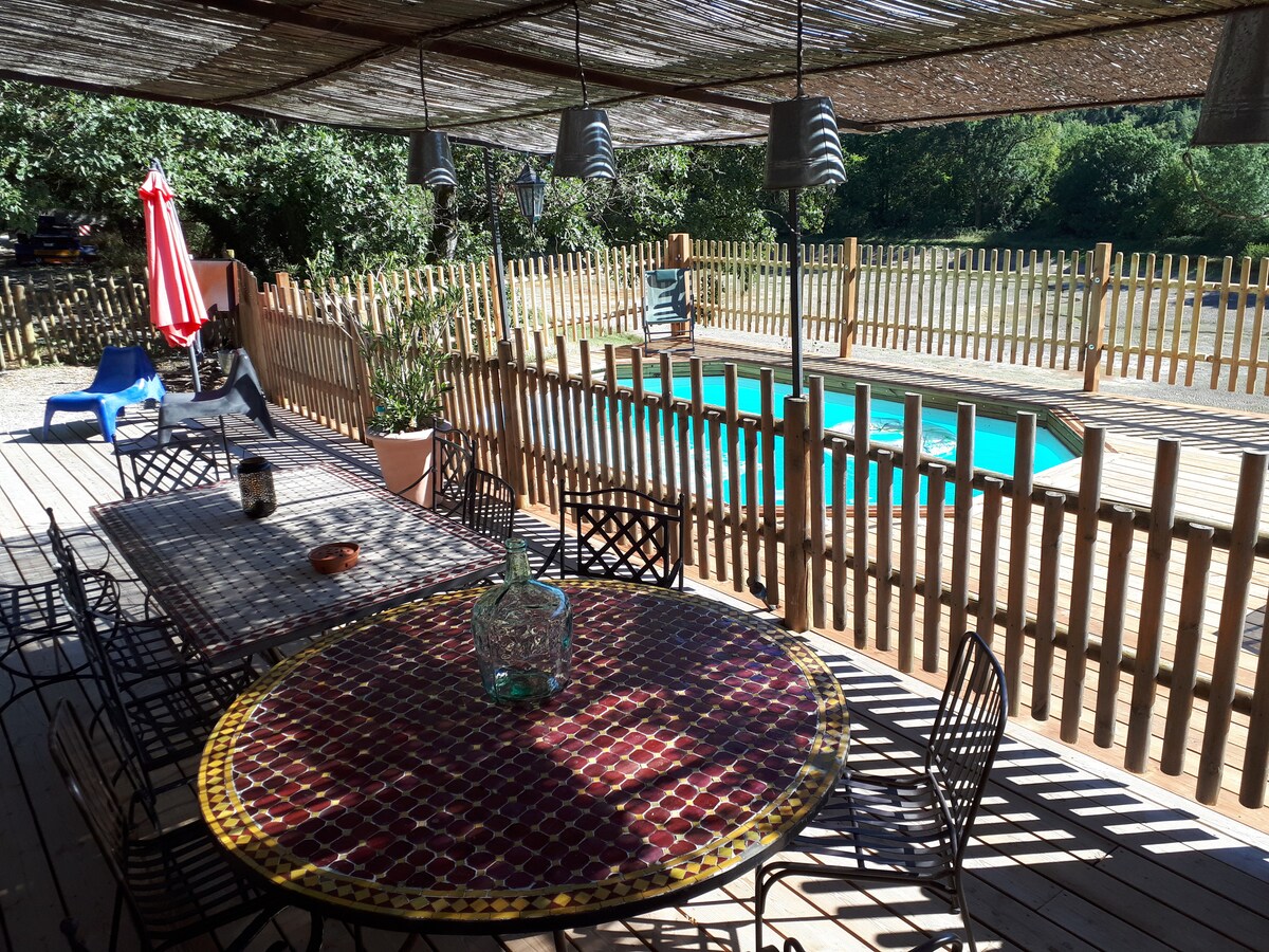An outdoor terrace is arranged with a large, round table surrounded by black wrought-iron chairs. A shaded area is created by a woven cover above. A private swimming pool is visible in the background, framed by a wooden fence and natural greenery.