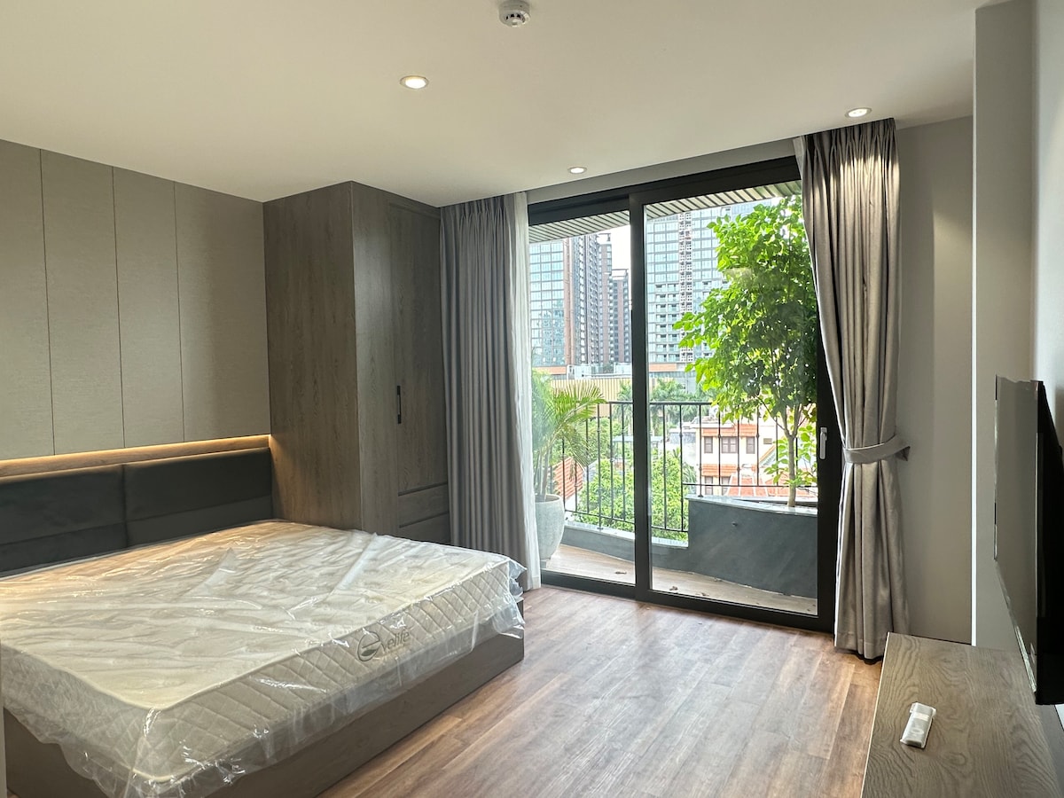 A spacious room features a large window with views of surrounding greenery and city buildings. The sleek wooden furniture includes a built-in wardrobe and a minimalist television console. A mattress is positioned on the bed frame, with the room bathed in natural light.