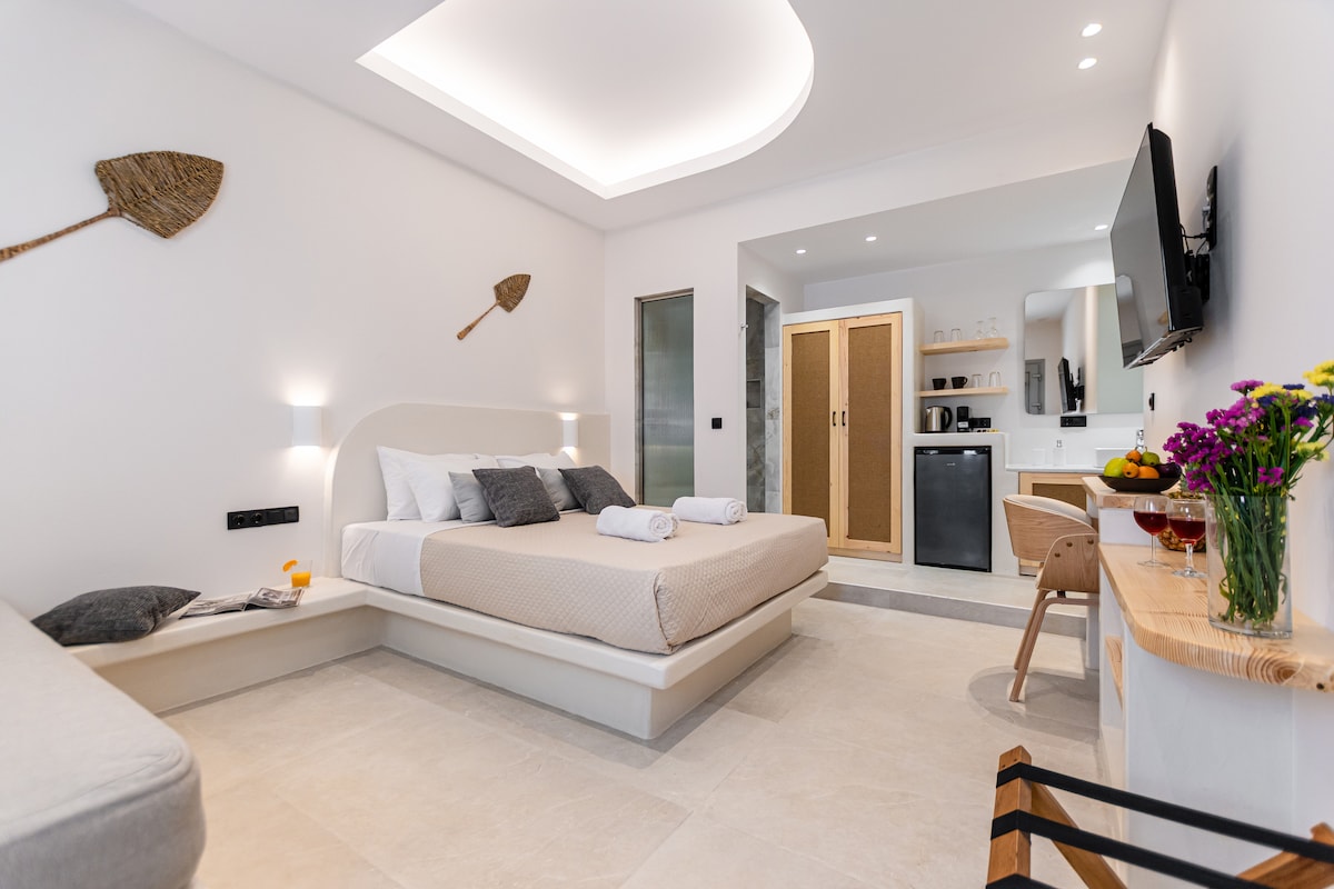 A spacious room features a sleek, elevated bed with neutral bedding and two folded towels. A small kitchenette area is visible with a refrigerator and coffee amenities. Stylish wall decor includes two decorative traditional brooms. Natural light illuminates the room through a modern ceiling design.