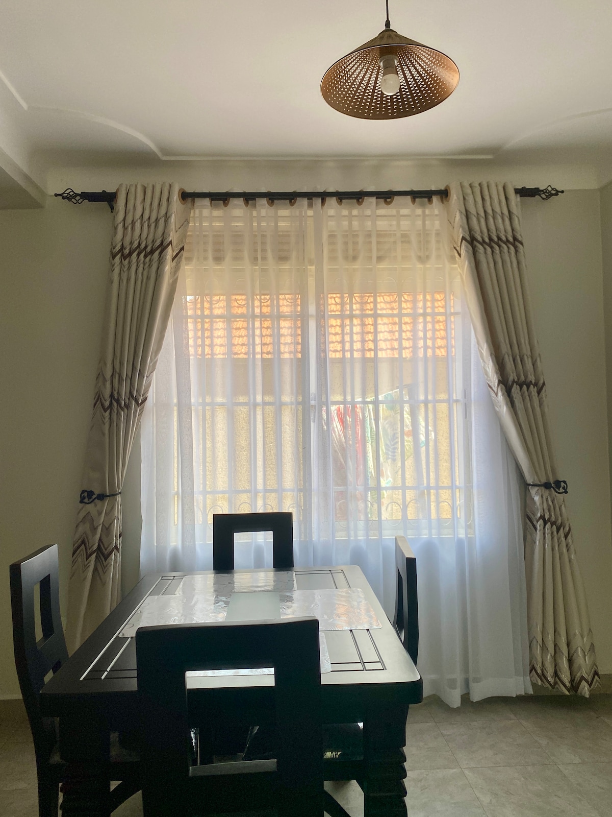 A dining area features a dark wooden table surrounded by four matching chairs. Large windows with light, airy curtains allow natural light to brighten the space, creating an inviting atmosphere for meals or gatherings.