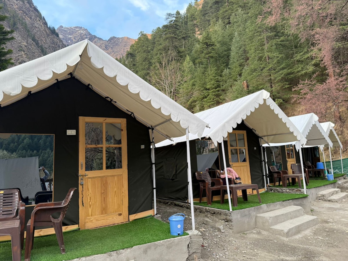 Two premium tents are viewed in the foreground, each featuring a wooden door and a covered porch area. Comfortable seating is arranged outside each tent, complemented by artificial grass. The backdrop highlights a mountainous landscape, with trees lining the site.