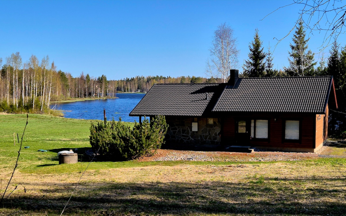A wooden cabin sits by a serene lake, surrounded by greenery. Large windows provide views of the water, while a pitched roof complements the natural setting. The landscape showcases a mix of grass and trees, promoting a sense of privacy and tranquility.